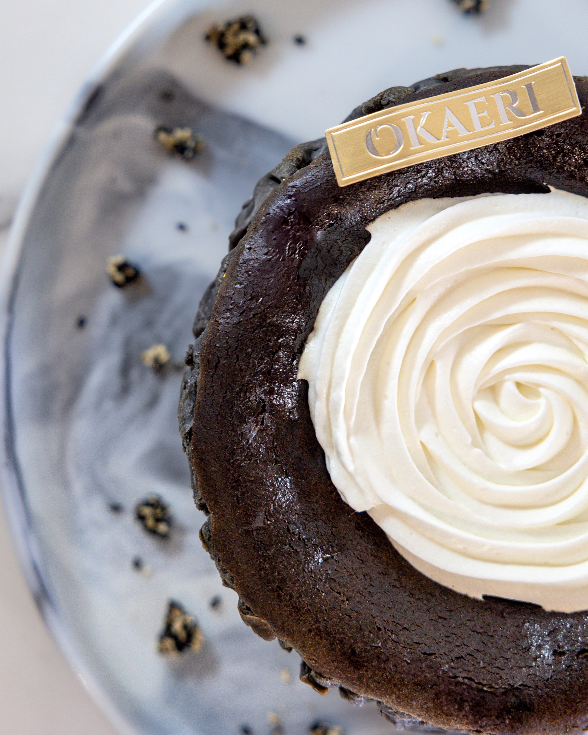 A slice of Kuki black sesame Basque burnt cheesecake with a swirl of white cream on top, garnished with a 'OKAERI' name tag and sesame brittles around the plate.
Okaeri Bakery - London based Asian Bakery