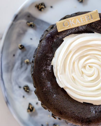 A slice of Kuki black sesame Basque burnt cheesecake with a swirl of white cream on top, garnished with a 'OKAERI' name tag and sesame brittles around the plate.
Okaeri Bakery - London based Asian Bakery