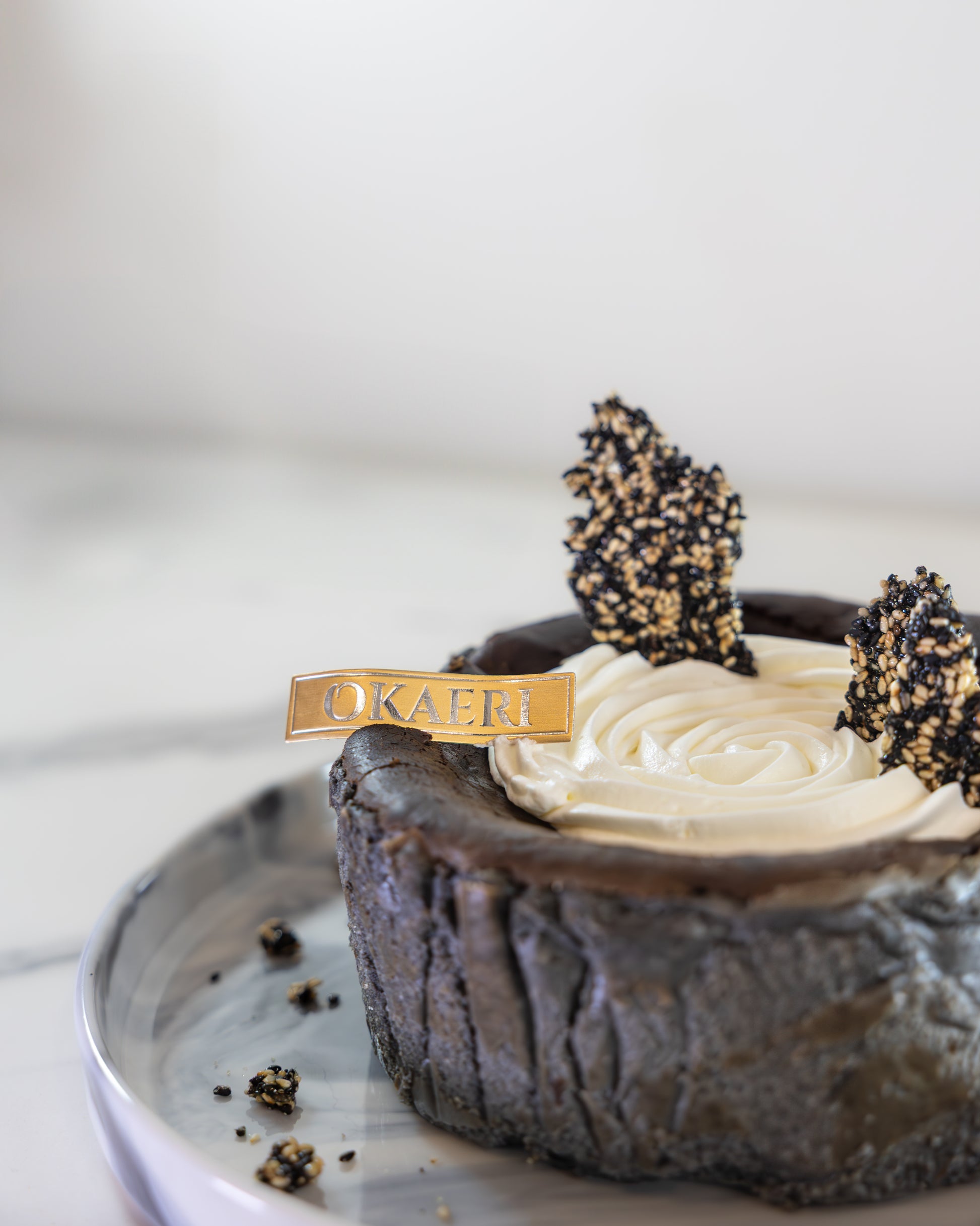 A slice of Kuki black sesame Basque burnt cheesecake with a swirl of white cream on top, garnished with a 'OKAERI' name tag and sesame brittles around the plate.
Okaeri Bakery - London based Asian Bakery
